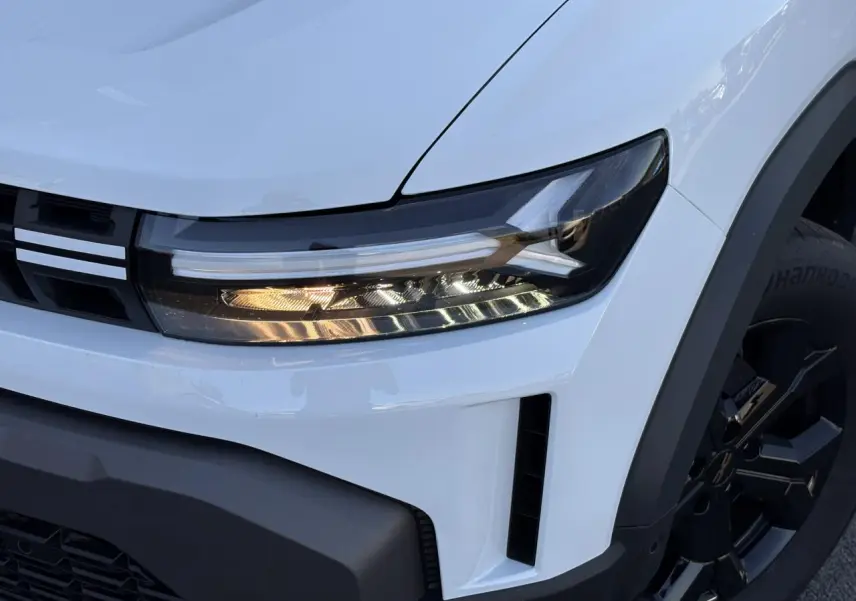Gros plan sur le phare avant droit et la calandre du Dacia Duster blanc Glacier avec jante alliage noire.
