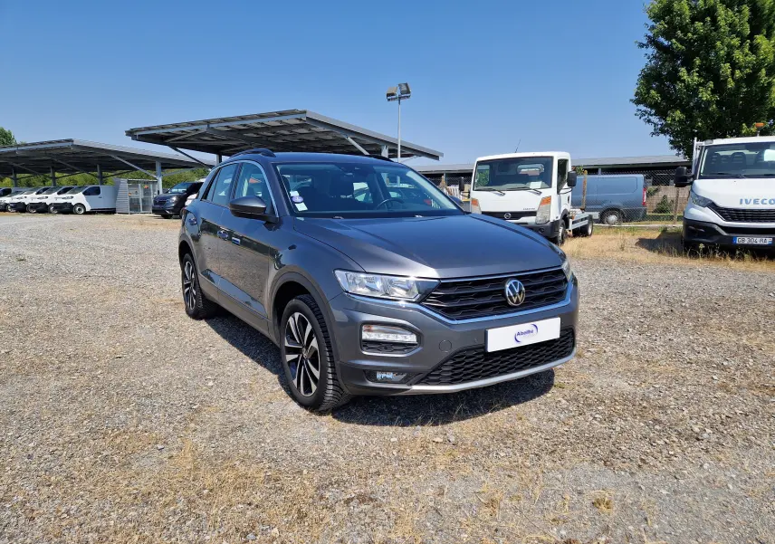 Volkswagen T-Roc gris foncé métal en 3/4 avant droit sur parking extérieur avec plusieurs véhicules utilitaires en arrière-plan.
