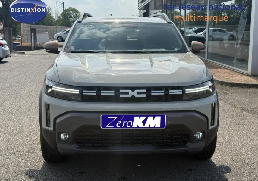 Vue avant d'un Dacia Duster blanc Glacier 2026 avec calandre noire et feux LED allumés sur parking extérieur.