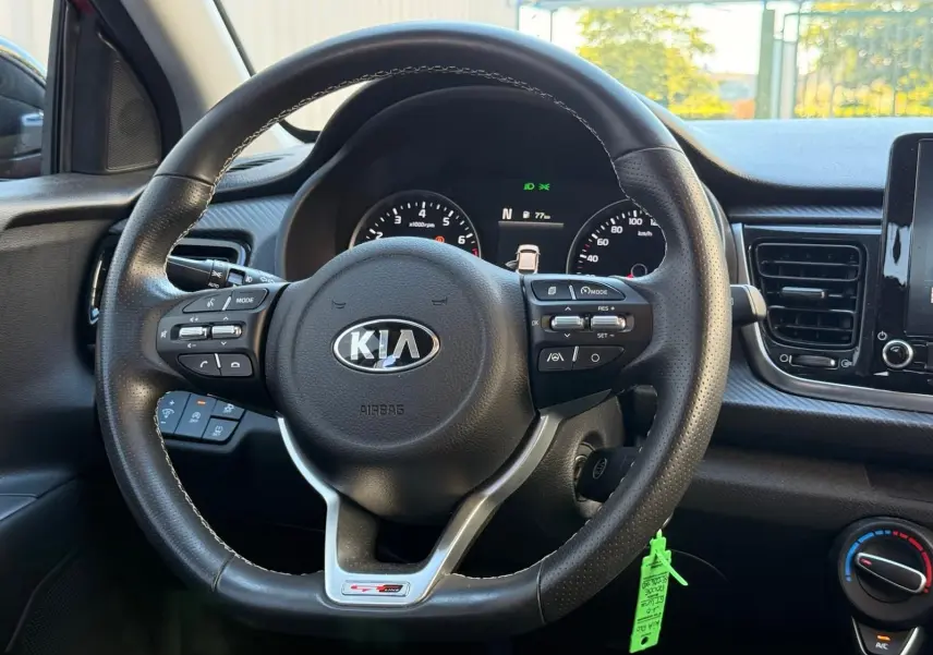 Vue intérieure centrée sur le volant en cuir de la KIA Rio rouge grenadine, avec commandes multifonctions et tableau de bord numérique.