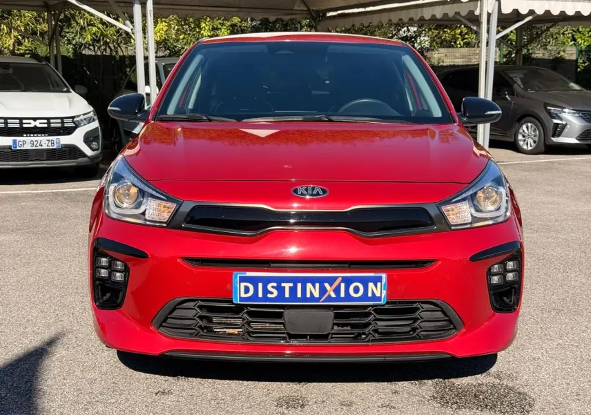 Vue frontale d'une KIA RIO rouge grenadine 2022 avec calandre noire et phares allumés, immatriculée Distinxion.
