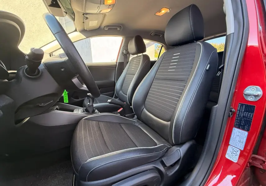 Intérieur côté conducteur d'une KIA Rio rouge grenadine 2022, sièges noirs avec surpiqûres blanches et volant visible.