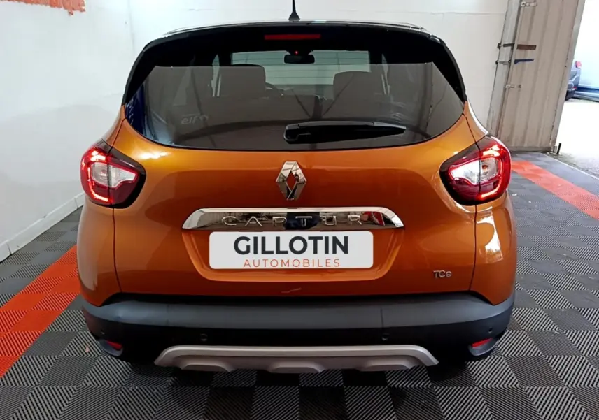 Vue arrière d'un Renault Captur orange avec feux allumés et logo chromé sur fond noir.