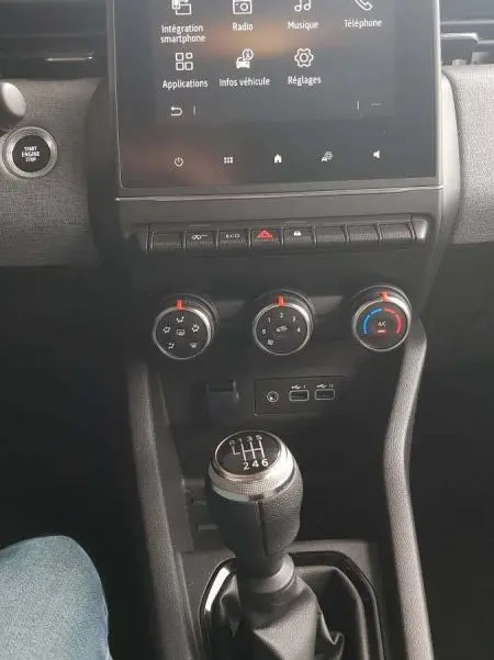 Vue intérieure centrée sur la console centrale d'une Renault Clio gris 2025, avec écran tactile et levier de vitesses manuel.