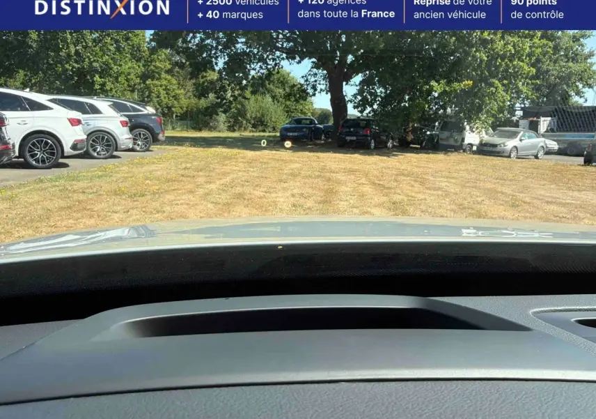 Vue depuis l'intérieur du Volvo XC90 gris 2024, montrant le tableau de bord et le pare-brise vers un parking extérieur.