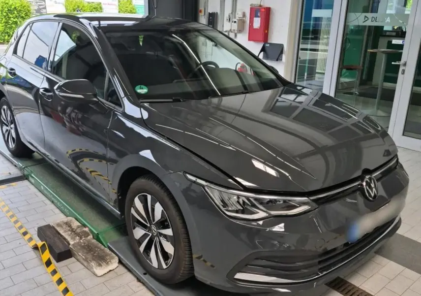 Volkswagen Golf 8 gris Urano vue 3/4 avant droit, avec jantes alliage et phares LED allumés en intérieur showroom.