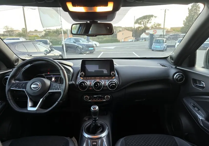 Intérieur noir du Nissan Juke II 2024 vu de face, avec volant cuir, écran tactile central et levier de vitesses manuel.