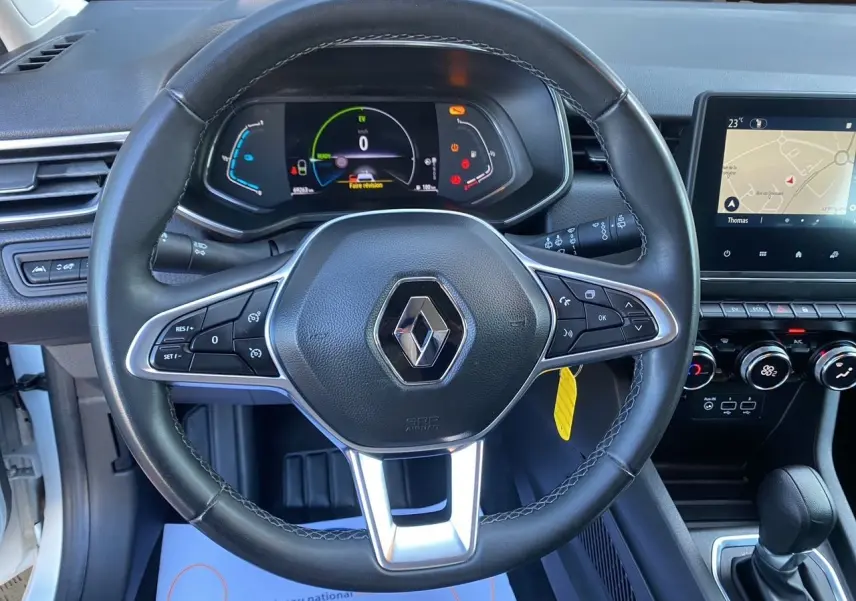 Vue rapprochée du volant en cuir noir avec logo Renault, tableau de bord numérique et écran tactile du Renault Clio V blanc.