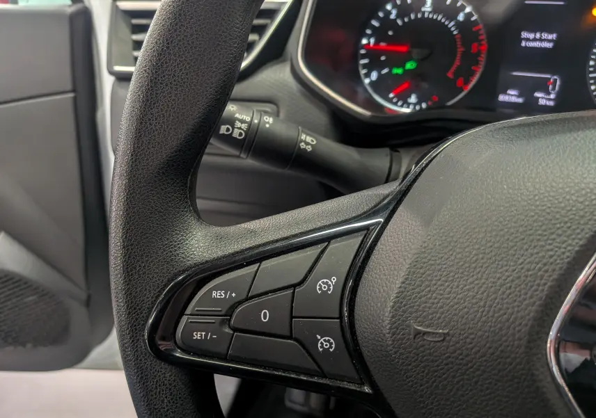 Gros plan sur le volant côté gauche de la Renault Clio V blanche, montrant les commandes de régulateur de vitesse et le tableau de bord flou.