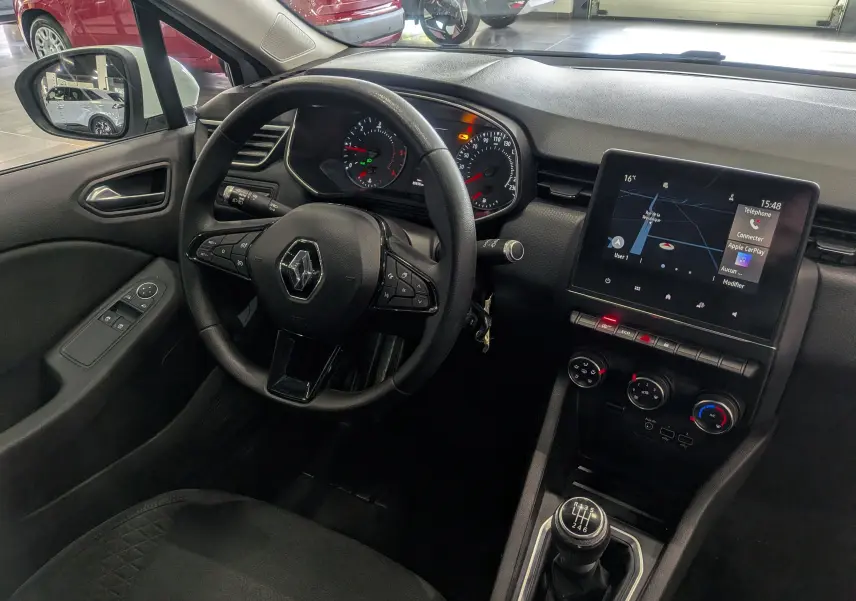 Vue intérieure côté conducteur de la Renault Clio V blanche, avec volant multifonction, écran tactile et levier de vitesse manuel.