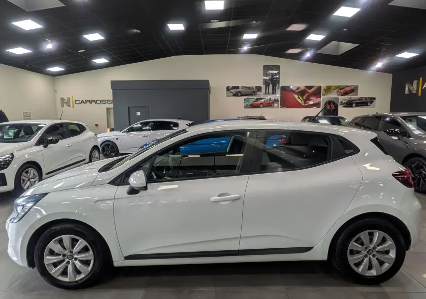 Vue de profil côté gauche d'une Renault Clio V blanche Glacier en intérieur de showroom, avec jantes alliage et vitres teintées.