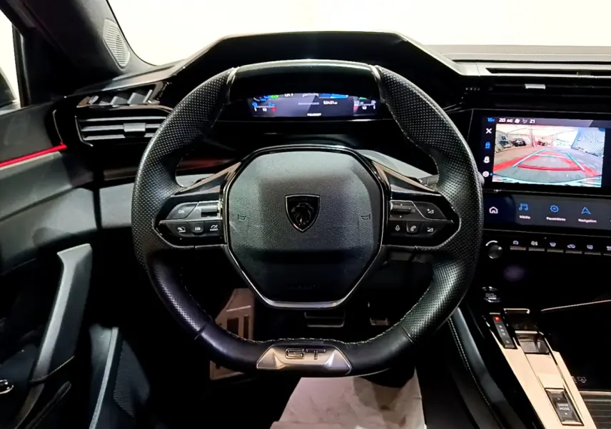 Vue intérieure centrée sur le volant cuir noir GT et le tableau de bord numérique de la Peugeot 308 PHEV 180 blanche.