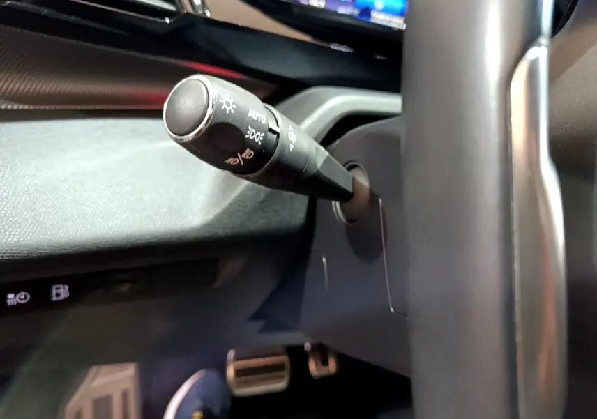 Gros plan sur la manette de commande d'éclairage à gauche du volant dans l'habitacle sombre d'une Peugeot 308 PHEV blanche.