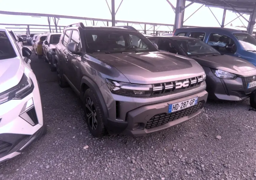 Vue 3/4 avant d'un Dacia Duster gris foncé garé sous abri, avec calandre et phares LED distinctifs visibles.