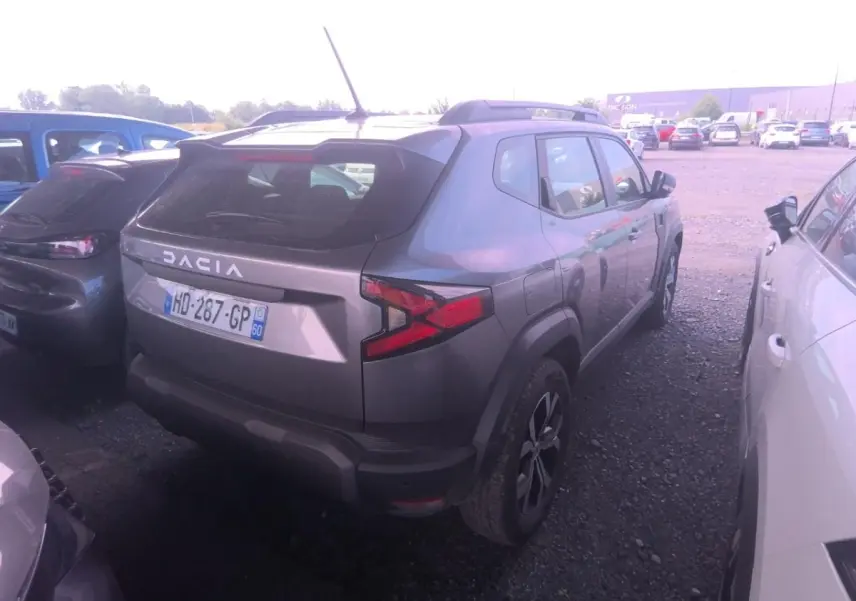 Vue 3/4 arrière droite d'un Dacia Duster gris foncé avec rails de toit noirs, stationné entre deux voitures.
