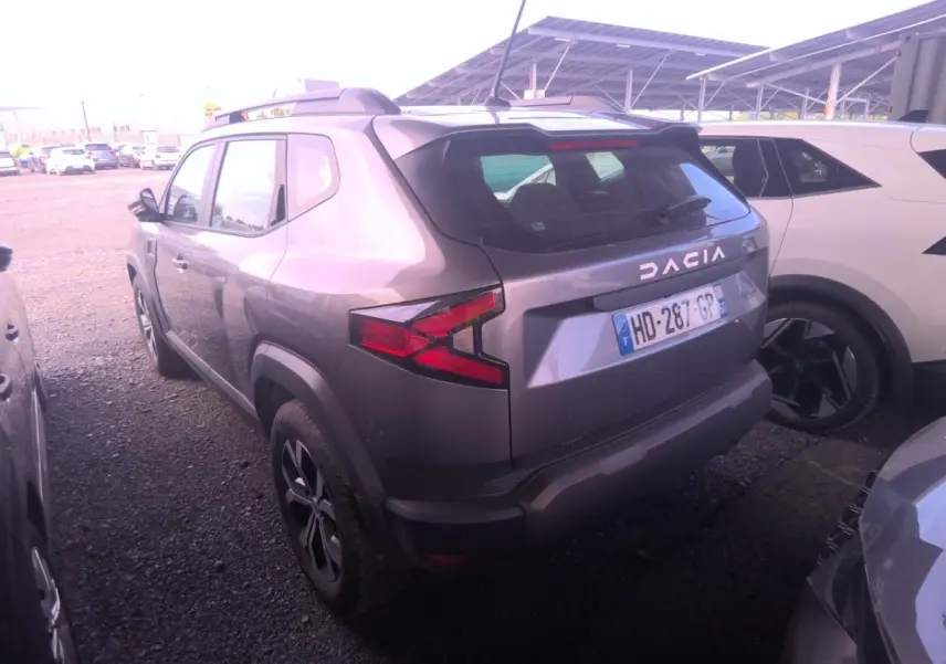 Vue 3/4 arrière d'un Dacia Duster gris foncé avec rails de toit noirs et feux arrière LED, stationné entre deux véhicules.