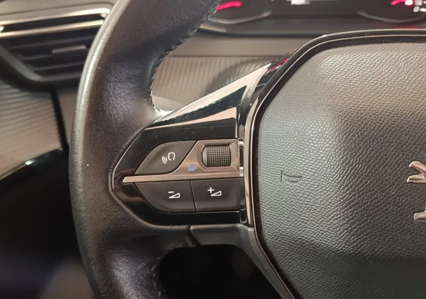 Gros plan sur le volant cuir noir de la Peugeot 208 blanc, avec commandes audio et bouton molette à gauche.