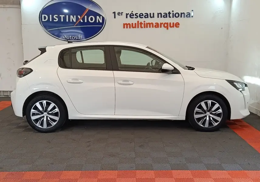 Profil droit d'une Peugeot 208 blanche PureTech 100 S&S Active Business, stationnée en intérieur sur sol damier noir et orange.