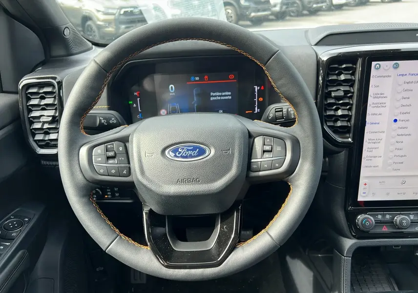 Vue centrée du volant en cuir noir avec surpiqûres orange du Ford Ranger 2025, tableau de bord numérique et écran tactile visible.