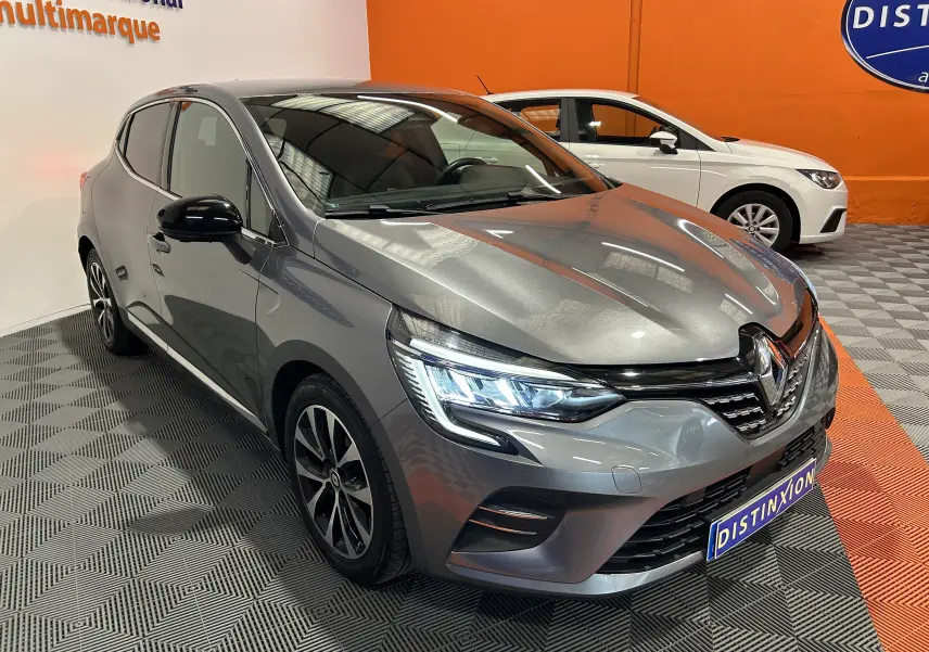 Vue 3/4 avant gauche d'une Renault Clio V gris métallisé 2023 avec phares LED allumés et caméra 360° visible sur le pare-chocs.