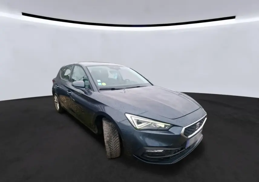 SEAT Leon gris 3/4 avant droit, phares allumés, roues légèrement tournées, sur fond neutre intérieur.