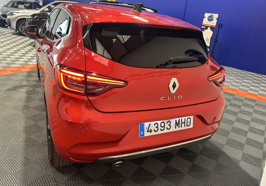 Vue 3/4 arrière d'une Renault Clio V rouge 2023 avec feux allumés dans un showroom intérieur.