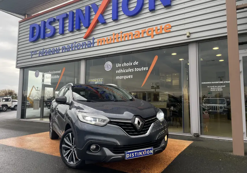 Renault Kadjar gris titanium en 3/4 avant droit devant un showroom Distinxion avec phares allumés et jantes alliage biton.