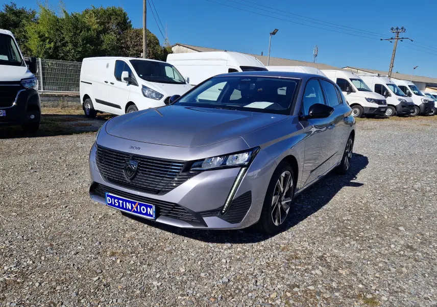 Peugeot 308 gris Artense vue 3/4 avant droit sur parking gravillonné avec plusieurs fourgons blancs en arrière-plan.