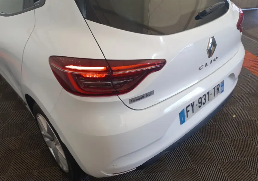 Vue 3/4 arrière droite d'une Renault Clio blanche avec feux arrière allumés et badge Renault Tech visible.