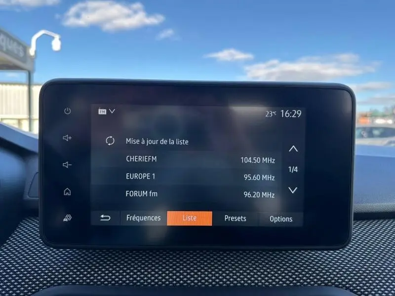 Écran tactile central du tableau de bord du Dacia Jogger 2025 affichant les stations radio FM, avec ciel bleu en arrière-plan.