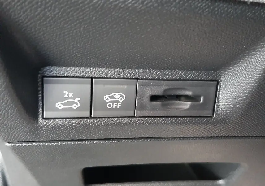 Gros plan sur les boutons de commande intérieurs noirs du Peugeot 3008 1.2 Hybrid bleu Ingaro, avec symboles d'ouverture de coffre et désactivation d'assistance.