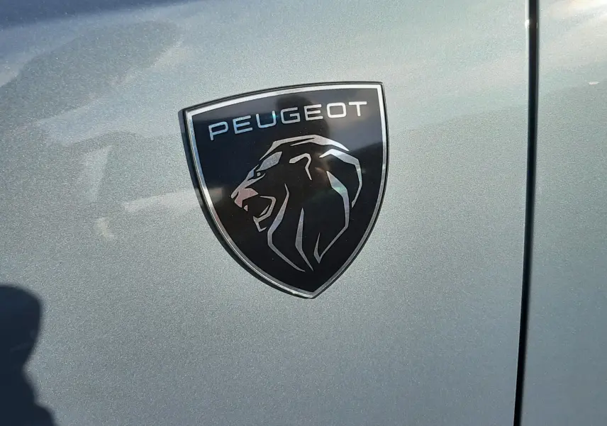 Gros plan sur l'écusson Peugeot noir et argent sur la carrosserie bleu Ingaro d'un 3008 hybride 2025.