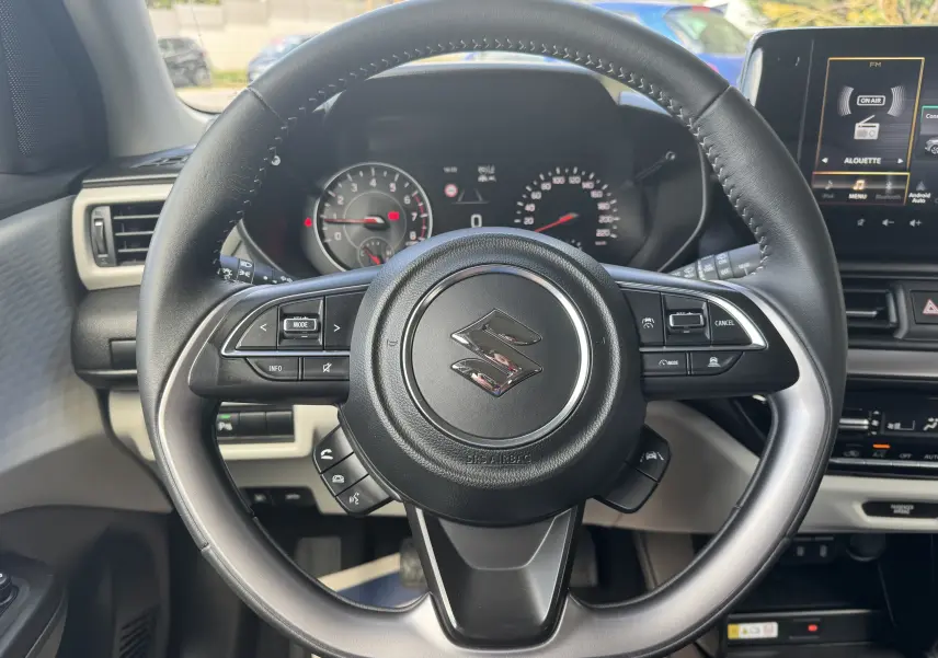 Vue centrée sur le volant noir de la Suzuki Swift 1.2 Pack Hybrid 2024 avec tableau de bord et écran multimédia en arrière-plan.