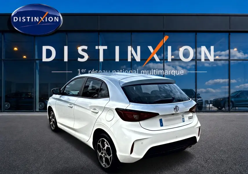 Vue 3/4 arrière droite d'une MG3 blanche 2026 hybride, avec vitres teintées et jantes alu devant un showroom Distinxion.