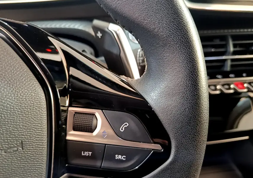 Gros plan sur les commandes gauche du volant cuir du Peugeot 2008 gris clair, avec tableau de bord en arrière-plan.