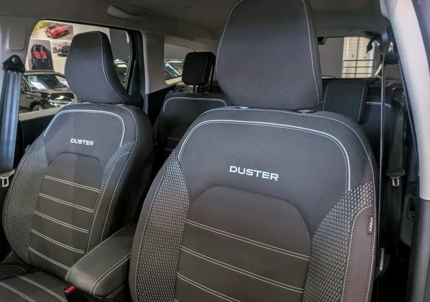 Gros plan sur les sièges avant en tissu noir avec surpiqûres blanches et logo DUSTER brodé, intérieur du Dacia Duster 2023.