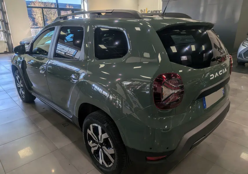 Vue 3/4 arrière droite du Dacia Duster 2023 kaki lichen avec jantes biton et barres de toit en showroom.