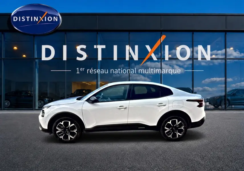 Vue de profil côté gauche d'une Citroën C4 X blanche 2025 avec jantes alu noires devant un bâtiment vitré.
