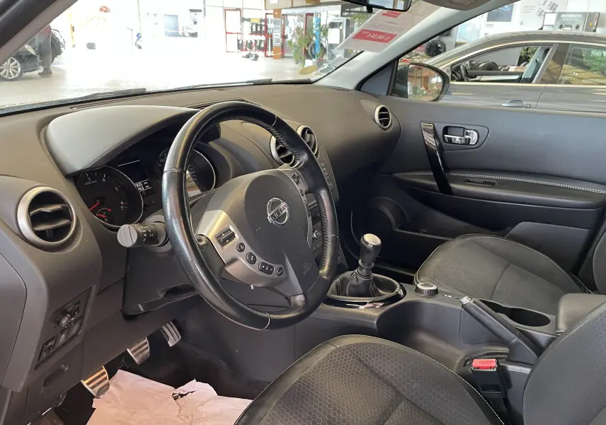 Intérieur noir du Nissan Qashqai 2013 vu côté conducteur, volant cuir, levier de vitesse manuel et tableau de bord visible.
