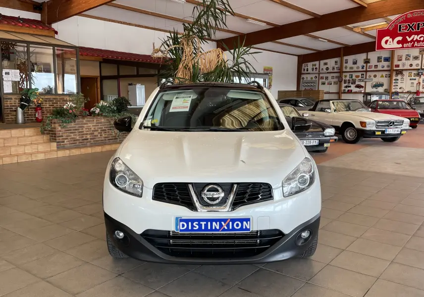 Nissan Qashqai blanc en vue frontale, intérieur showroom avec plusieurs voitures classiques en arrière-plan.