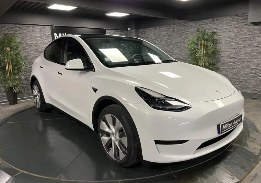 Tesla Model Y blanc nacré vu en 3/4 avant droit dans un showroom avec toit panoramique en verre visible.