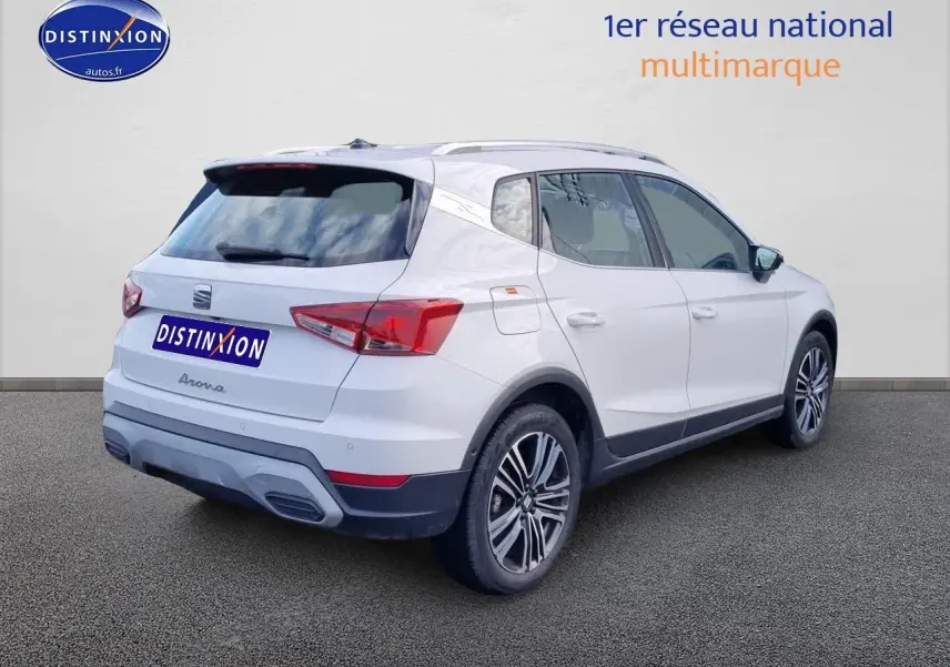 Vue 3/4 arrière droite d’un SEAT Arona blanc avec jantes alu et barres de toit sur fond neutre.