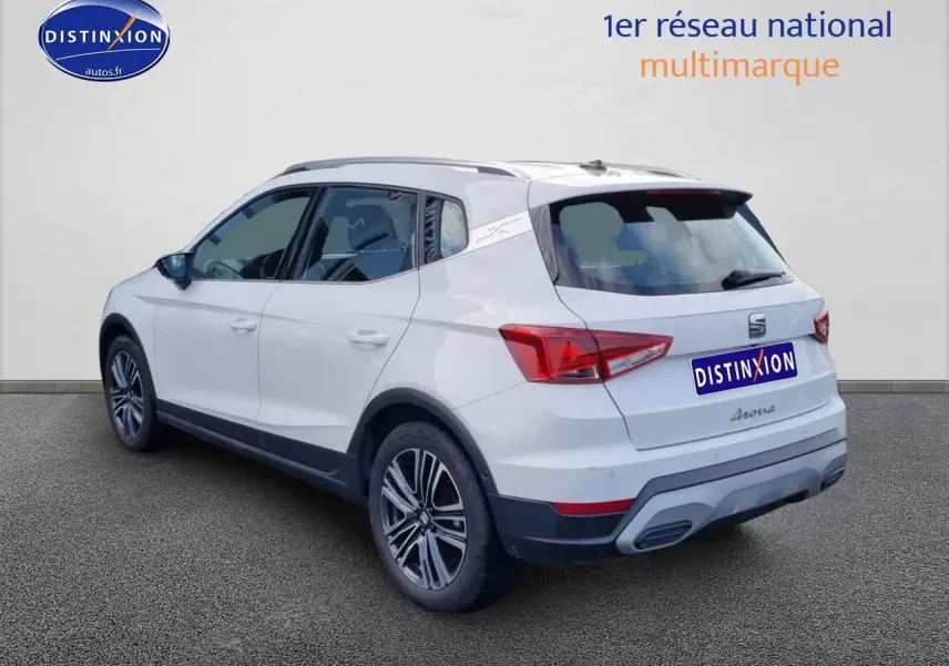 Vue 3/4 arrière droite d'un SEAT Arona blanc 2023 avec jantes alu et barres de toit visibles.