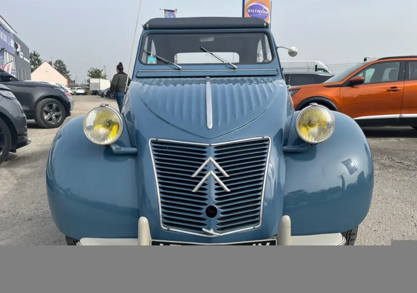 Vue frontale d'une Citroën 2 CV bleu clair de 1960 avec phares ronds jaunes et calandre à chevrons emblématiques.