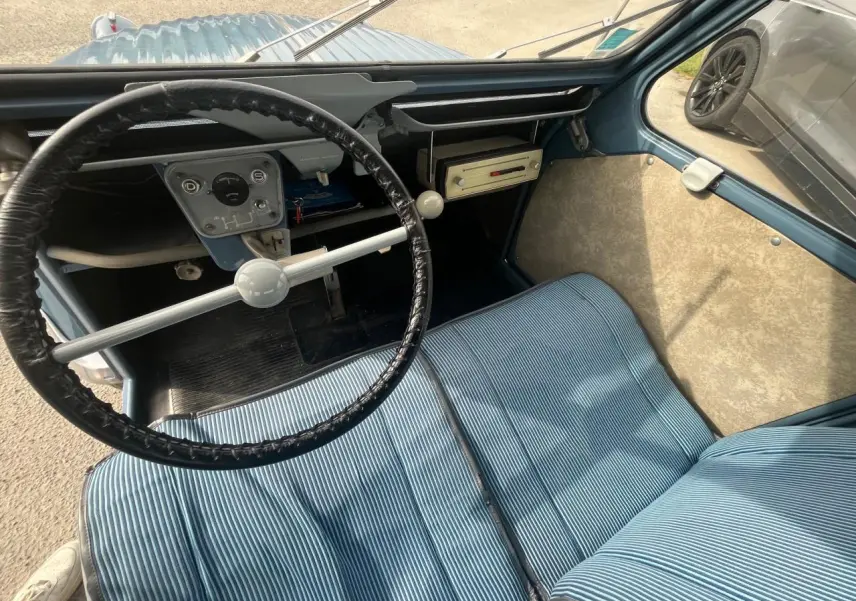 Vue intérieure du poste de conduite d'une Citroën 2 CV 1960 bleu clair, avec volant fin et sièges rayés bleus.