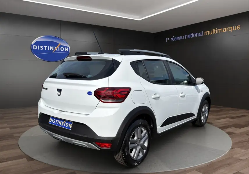 Vue 3/4 arrière droite d'une Dacia Sandero Stepway blanche avec barres de toit et protections noires visibles.