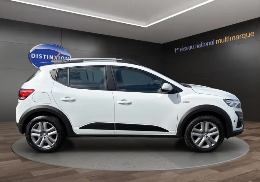 Profil droit de la Dacia Sandero Stepway blanche 2022 avec protections noires et barres de toit visibles en intérieur showroom.
