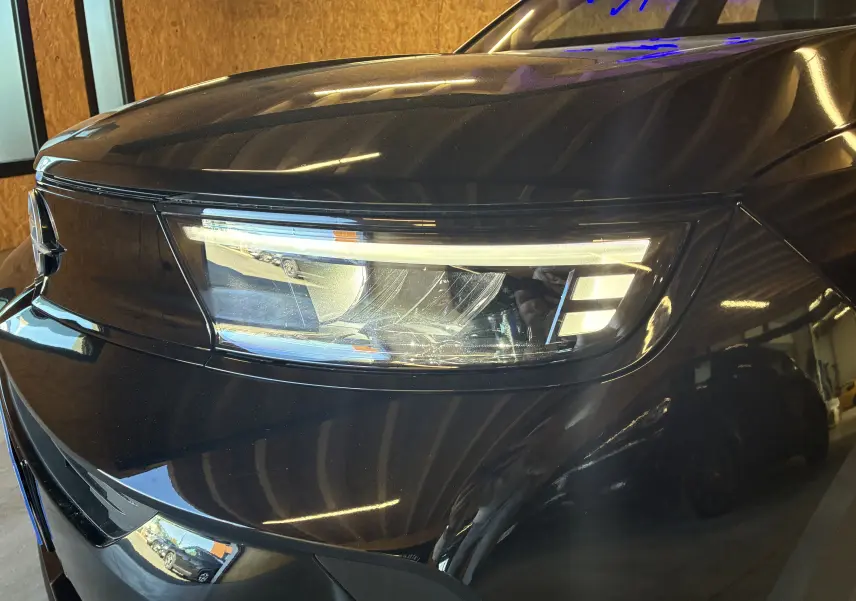 Gros plan sur le phare avant gauche noir brillant de l’Opel Mokka 1.2 Turbo Hybrid 2025 en intérieur.