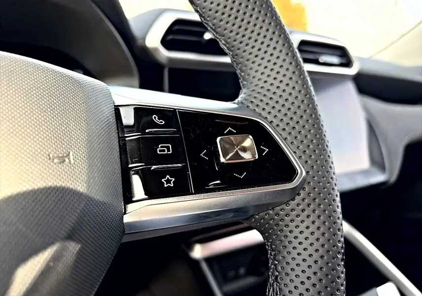 Gros plan sur les commandes au volant perforé du MG Motor ZS 1.5 hybrid 197 Cosmic Silver, tableau de bord en arrière-plan.