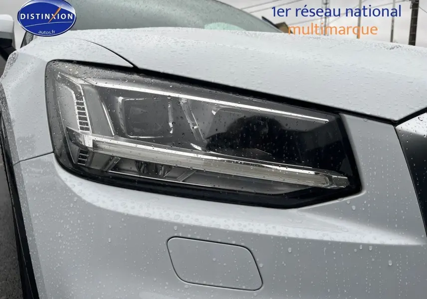 Gros plan sur le phare avant droit d’un Audi Q2 blanc Ibis avec gouttes de pluie visibles sur la carrosserie.